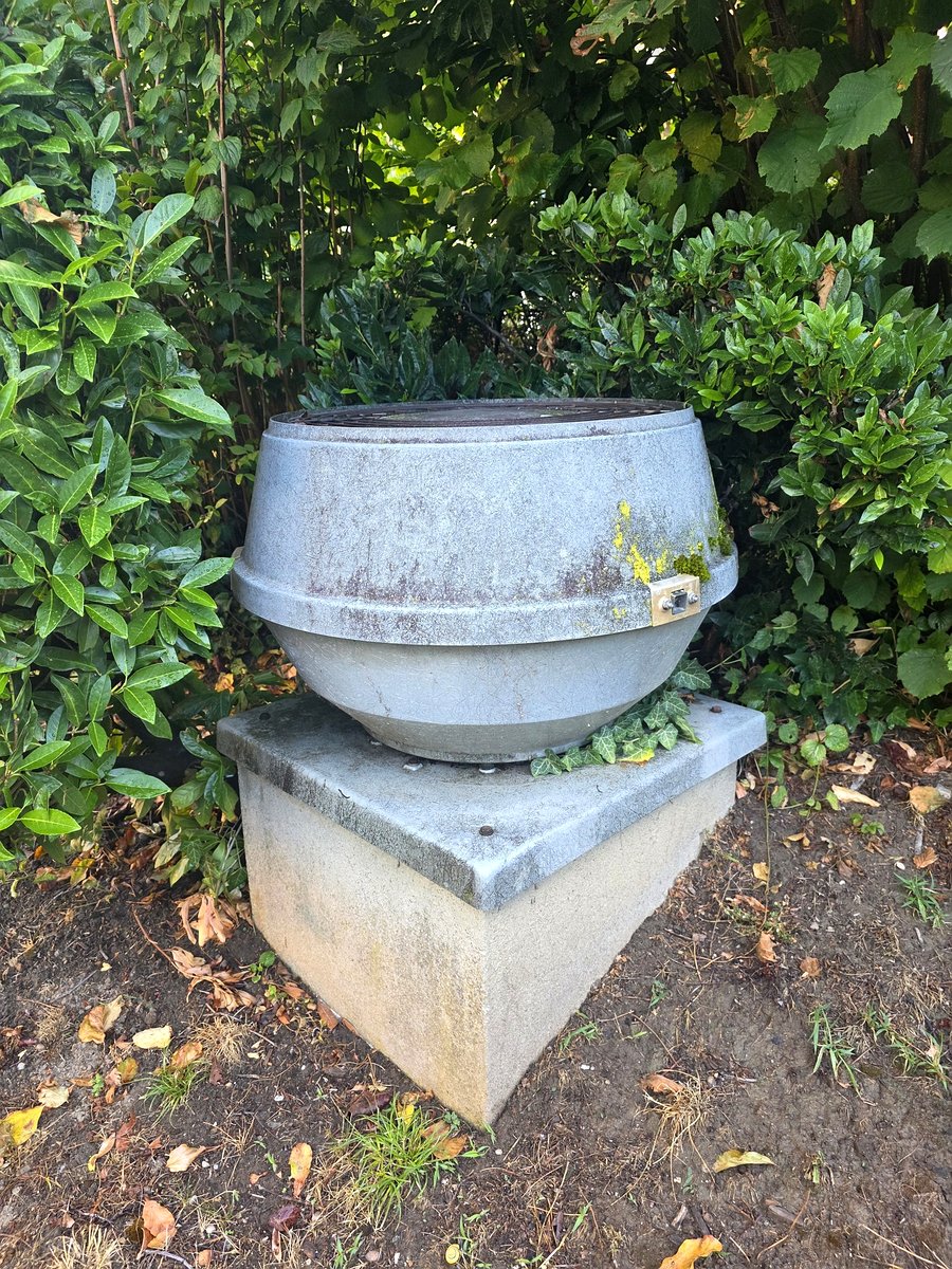 Le rejet d’air posé sur son socle en béton, au ras du sol, dans un massif planté — la haie vient toucher le capot. À quelques mètres, les fenêtres des voisins qui se plaignent.